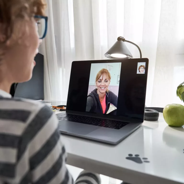 Back view of crop kid in glasses sitting at table with laptop and talking on video call with teacher while having online lesson during remote studies at home